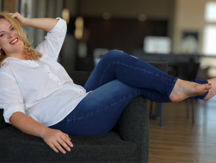 Woman reclining on a chair in a white blouse and blue jeans, smiling confidently in a casual, relaxed style.