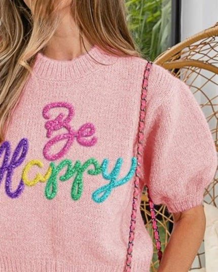 Person wearing a pink sweater with 'Be Happy' text in colorful letters, sitting on a wicker chair.