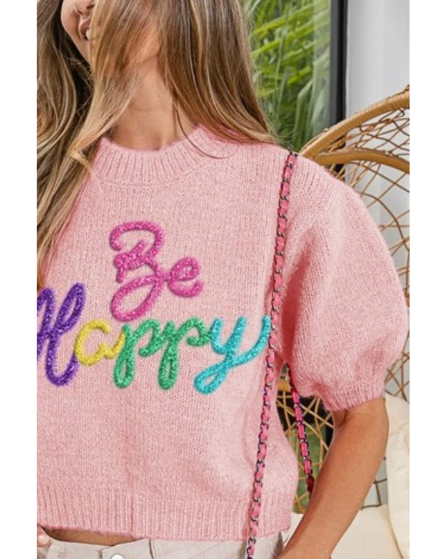 Person wearing a pink sweater with 'Be Happy' text in colorful letters, sitting on a wicker chair.
