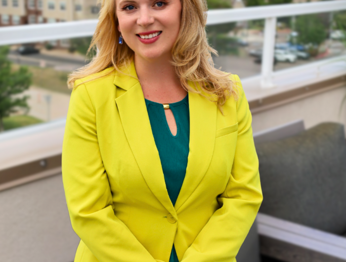 Woman in a lime green blazer and blue jeans standing on a balcony with a cityscape background