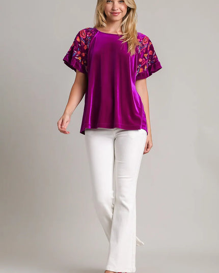 Woman wearing a purple blouse with floral sleeves and white pants on a plain background