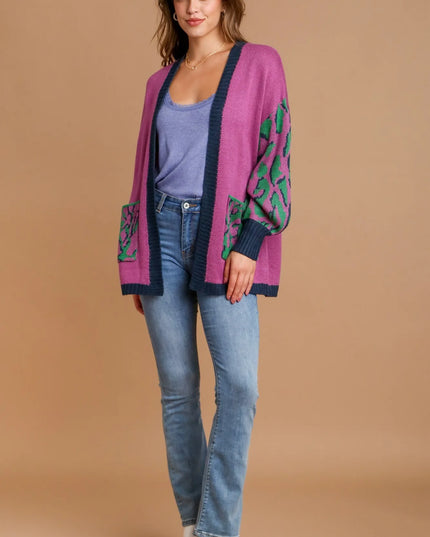 Woman wearing a pink cardigan with green patterns, blue jeans, and white shoes on a beige background.