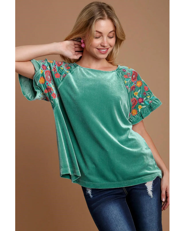 Woman wearing a teal/mint top with floral embroidery on sleeves against a brown background