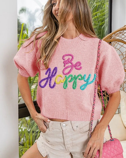 Woman wearing a pink sweater with 'Be Happy' text, standing outdoors.